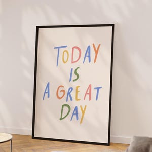 May include: A framed poster with the words "TODAY IS A GREAT DAY" in colorful, handwritten-style lettering. The poster has a black frame and is leaning against a white wall. The text is in blue, yellow, green, orange, and pink.