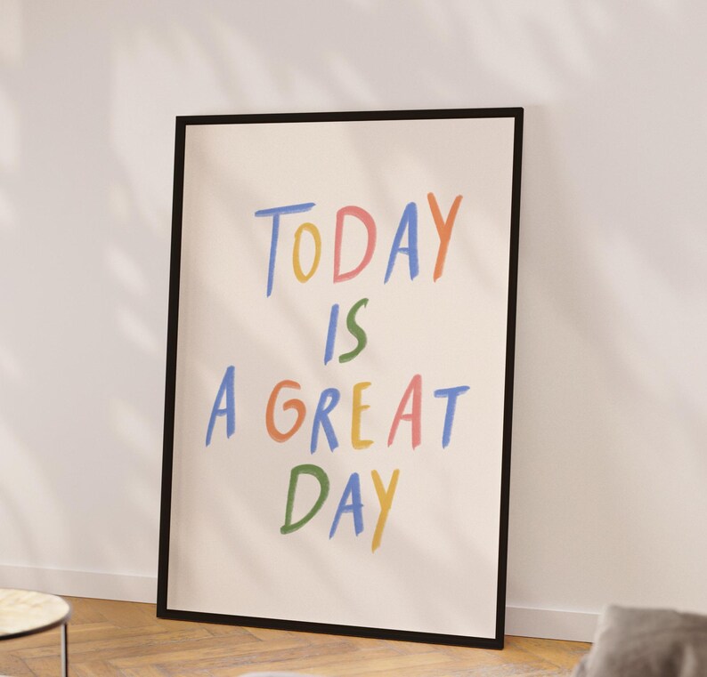 May include: A framed poster with the words "TODAY IS A GREAT DAY" in colorful, handwritten-style lettering. The poster has a black frame and is leaning against a white wall. The text is in blue, yellow, green, orange, and pink.