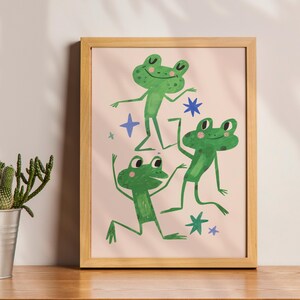 May include: A framed art print featuring three green frogs in playful poses against a light pink background. The frogs are depicted with simple, whimsical details, surrounded by blue star shapes. The frame is a light wood color.
