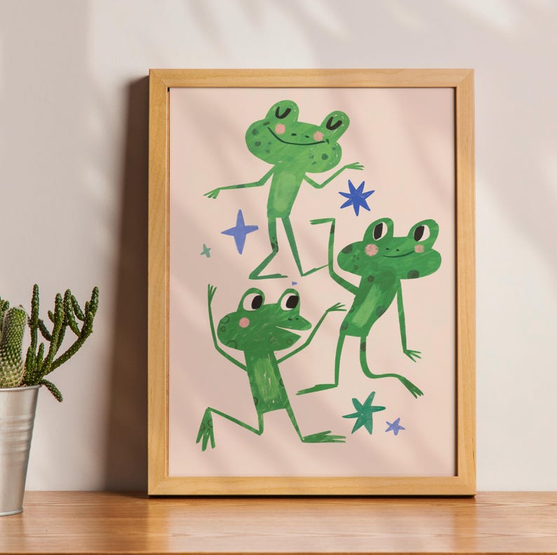 May include: A framed art print featuring three green frogs in playful poses against a light pink background. The frogs are depicted with simple, whimsical details, surrounded by blue star shapes. The frame is a light wood color.