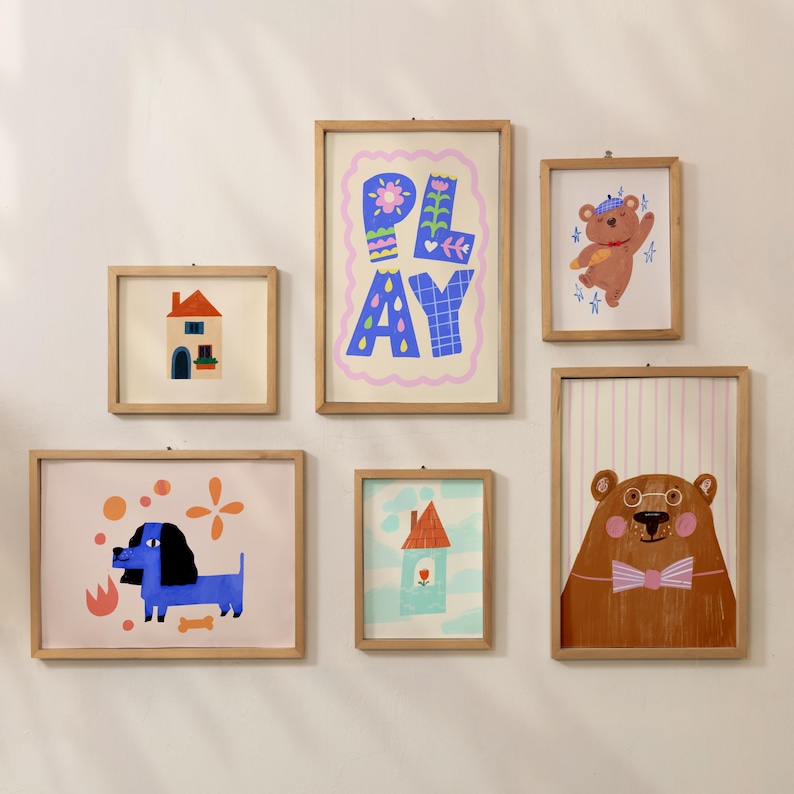 K&ouml;nnte beinhalten: Eine Sammlung von sechs gerahmten Kunstdrucken mit skurrilen Illustrationen. Die Drucke zeigen ein Haus, einen Hund, einen B&auml;ren und das Wort "PLAY" in einem farbenfrohen Design. Die Rahmen sind aus hellem Holz.