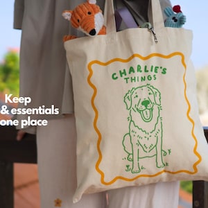 May include: A cream-colored canvas tote bag featuring a green line-art illustration of a dog and the text "CHARLIE'S THINGS" enclosed in a wavy orange border. The bag contains a small orange fox plush toy and a blue plush toy inside the main compartment.