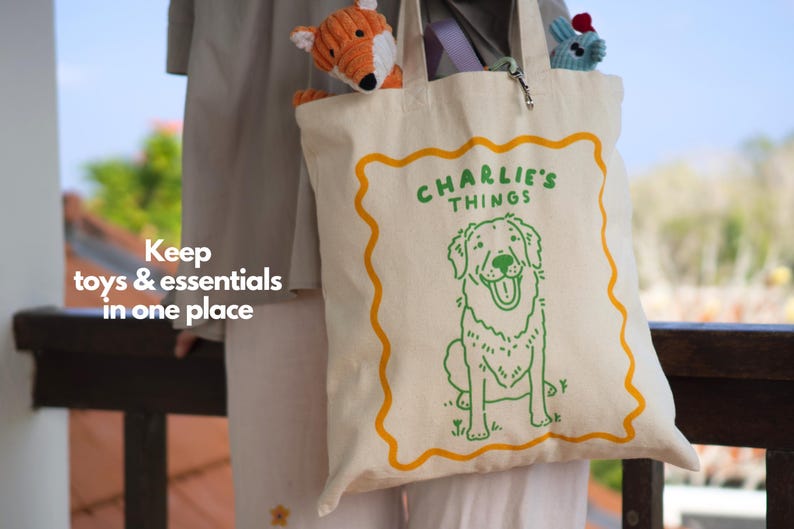 May include: A cream-colored canvas tote bag featuring a green line-art illustration of a dog and the text "CHARLIE'S THINGS" enclosed in a wavy orange border. The bag contains a small orange fox plush toy and a blue plush toy inside the main compartment.