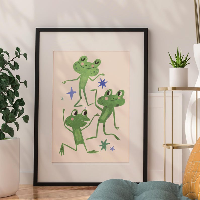 May include: A framed art print featuring three green cartoon frogs dancing on a light pink background. The frogs have big eyes and are surrounded by blue star shapes. The frame is black.