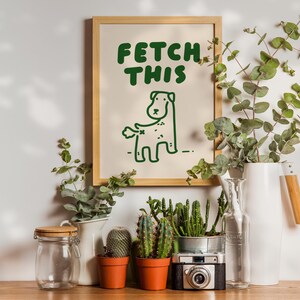 May include: A framed print with a green illustration of a dog with the text "FETCH THIS" on a beige background. The print is surrounded by potted plants and a vase with greenery.