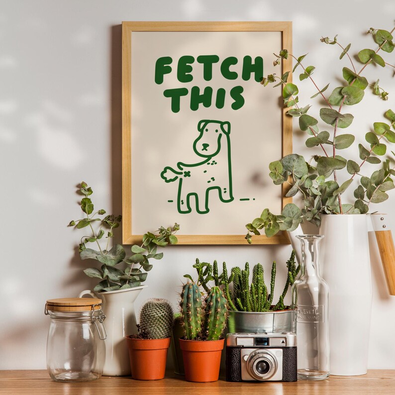 May include: A framed print with a green illustration of a dog with the text "FETCH THIS" on a beige background. The print is surrounded by potted plants and a vase with greenery.