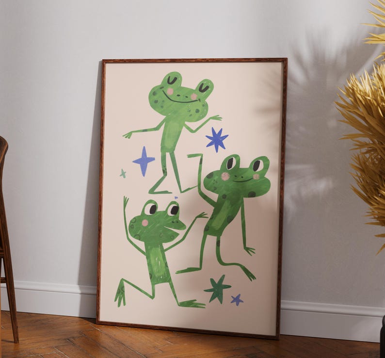 May include: A framed art print featuring three green cartoon frogs in various poses, with blue star accents on a cream background. The artwork has a whimsical, playful style, perfect for a child's room or a fun-loving space.
