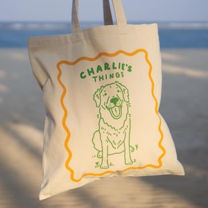 Op de afbeelding: Een crèmekleurige canvas draagtas met een feloranje rand van golvende lijnen. In het midden staat een groene lijntekening van een zittende hond, met de groene tekst "CHARLIE'S THINGS" boven de kop van de hond gedrukt.