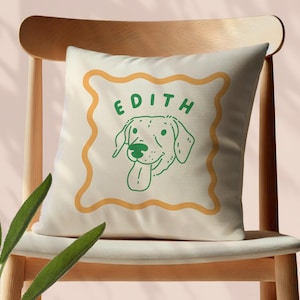 Pode incluir: Almofada quadrada creme com um design verde e amarelo. O design apresenta uma ilustração de cão e o nome "EDITH" em letras verdes. A almofada está numa cadeira de madeira.