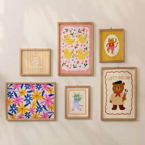 May include: A collection of framed art prints with whimsical illustrations. The prints feature a variety of designs, including floral patterns, cartoon animals, and the text "You are my sunshine". The frames are made of light-colored wood.
