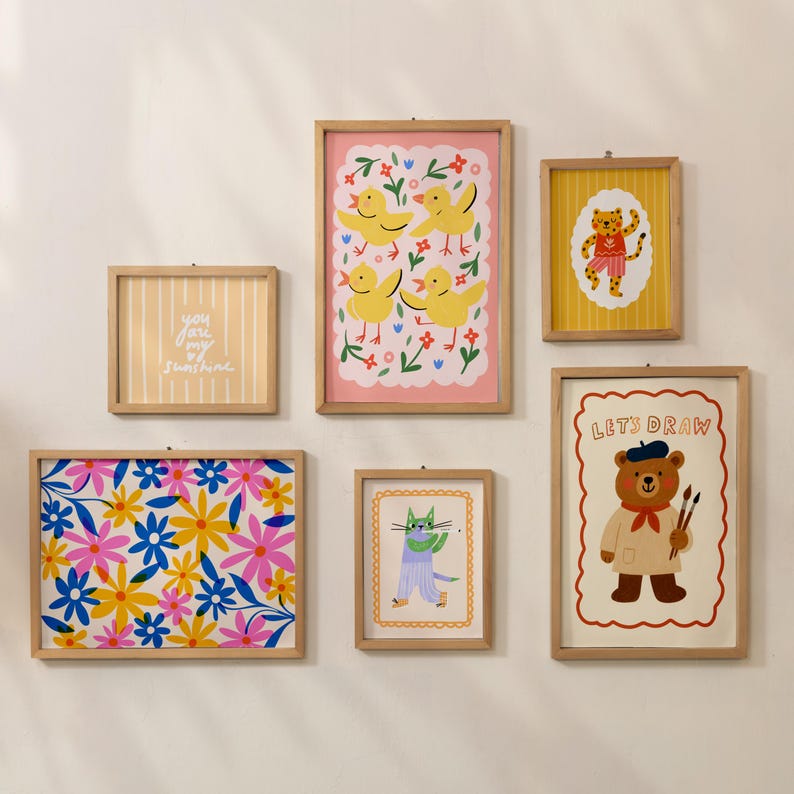 May include: A collection of framed art prints with whimsical illustrations. The prints feature a variety of designs, including floral patterns, cartoon animals, and the text "You are my sunshine". The frames are made of light-colored wood.