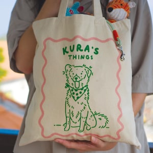 Op de afbeelding: Een beige canvas draagtas met een groene lijntekening van een hond met een bandana met stippen. Boven de hond staat de tekst KURA'S THINGS in het groen. Een roze golvende rand omlijst het ontwerp en er is een lichtgroene hondenriem aan bevestigd.