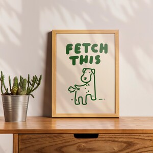 May include: A framed print with a light brown frame and a white background. The print features a green cartoon dog with the text "FETCH THIS" in green letters.