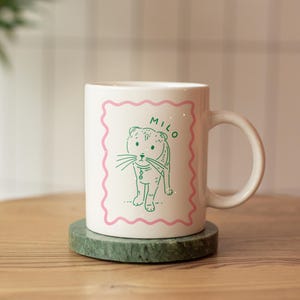 May include: White ceramic mug featuring a green line drawing of a cat with the name "Milo" in green text on a pink background.