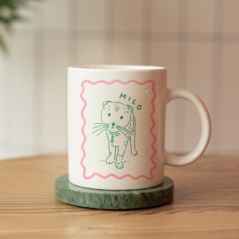 May include: White ceramic mug featuring a green line drawing of a cat with the name "Milo" in green text on a pink background.