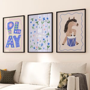May include: Three framed art prints with black frames. The left print says "PLAY" in colorful block letters. The middle print says "BE KIND" surrounded by flowers. The right print features a horse playing a drum.