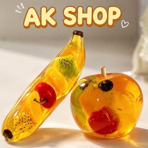 May include: A clear yellow resin banana and apple sculpture containing embedded fruit slices including kiwi, cherry, and dragon fruit. The text AK SHOP is displayed in large, light-coloured letters at the top of the image with a small heart icon.