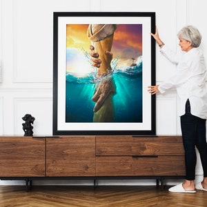 May include: A framed print of a painting depicting a hand reaching out from the water to grasp another hand. The painting is set against a backdrop of a sunset sky and ocean waves.