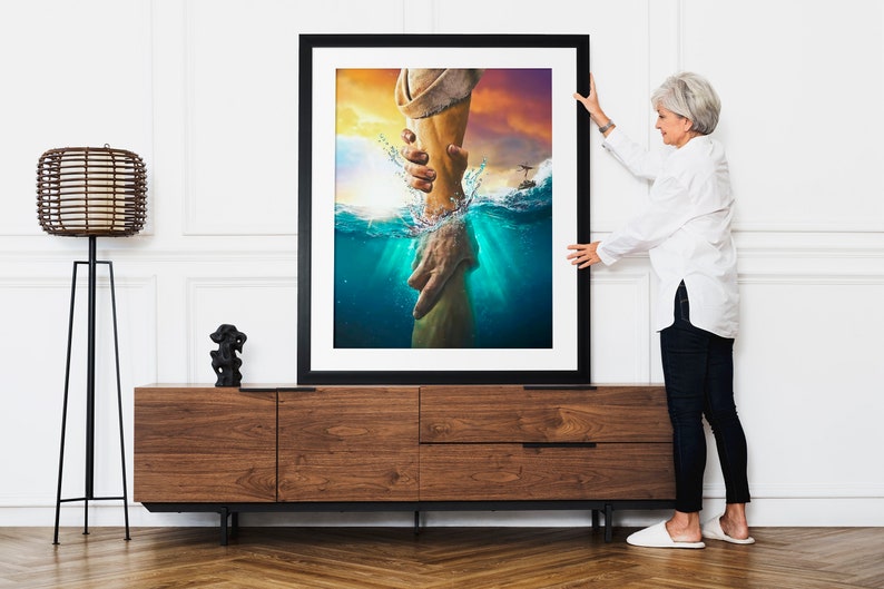 May include: A framed print of a painting depicting a hand reaching out from the water to grasp another hand. The painting is set against a backdrop of a sunset sky and ocean waves.