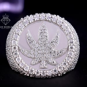 Puede incluir: Un anillo plateado con un diseño de hoja de cannabis. El anillo presenta una cara redonda con un motivo de hoja central incrustado con pequeñas piedras brillantes. La banda y el borde exterior de la cara también están adornados con piedras similares.