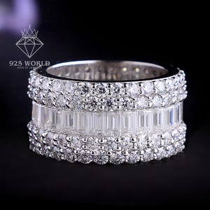 May include: A wide silver-toned band ring featuring a central row of rectangular baguette-cut clear stones flanked by two rows of small round brilliant-cut clear stones. The ring displays the text 925 WORLD above the band on a black background.