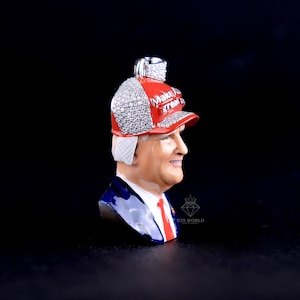 May include: A pendant featuring a bust of a man wearing a red baseball cap with the text "Make America Great Again". The cap is encrusted with sparkling stones. The man is wearing a blue suit with a red tie.