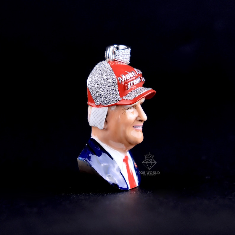 May include: A pendant featuring a bust of a man wearing a red baseball cap with the text "Make America Great Again". The cap is encrusted with sparkling stones. The man is wearing a blue suit with a red tie.