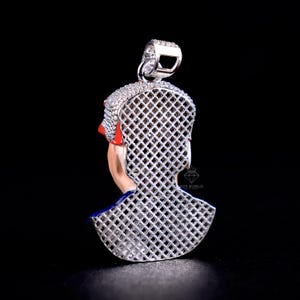 May include: A silver pendant with a detailed design of a person's profile. The pendant features a red and blue accent, with a lattice-like pattern on the back. The top of the pendant is encrusted with small, sparkling stones.