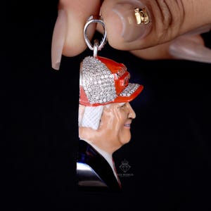 May include: A silver pendant featuring a portrait of a person wearing a red hat with a diamond-studded brim. The pendant has a detailed painted face and a black suit. The pendant is held by a silver loop.