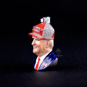 May include: A pendant featuring a bust of a man wearing a red baseball cap with the text "America Again". The cap is embellished with sparkling stones. The man is wearing a blue suit with a red tie and an American flag pin.