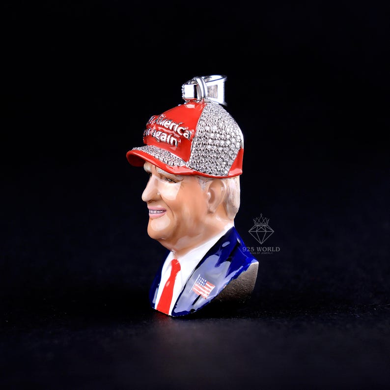 May include: A pendant featuring a bust of a man wearing a red baseball cap with the text "America Again". The cap is embellished with sparkling stones. The man is wearing a blue suit with a red tie and an American flag pin.