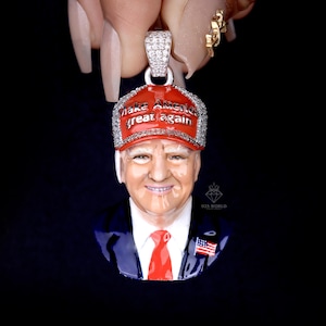 May include: A silver pendant featuring a detailed portrait of a man wearing a red baseball cap with the text "Make America Great Again". The pendant has a red tie and a small American flag pin.