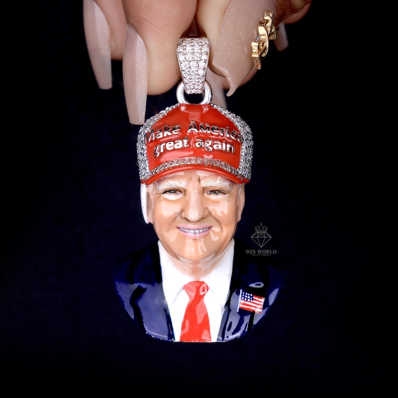 May include: A silver pendant featuring a detailed portrait of a man wearing a red baseball cap with the text "Make America Great Again". The pendant has a red tie and a small American flag pin.