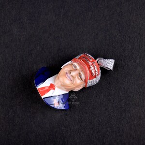 May include: A pendant featuring a smiling face with a red and white head covering that reads "Make America Great Again." The pendant has a silver bail and is set against a black background. The face is painted with realistic skin tones.