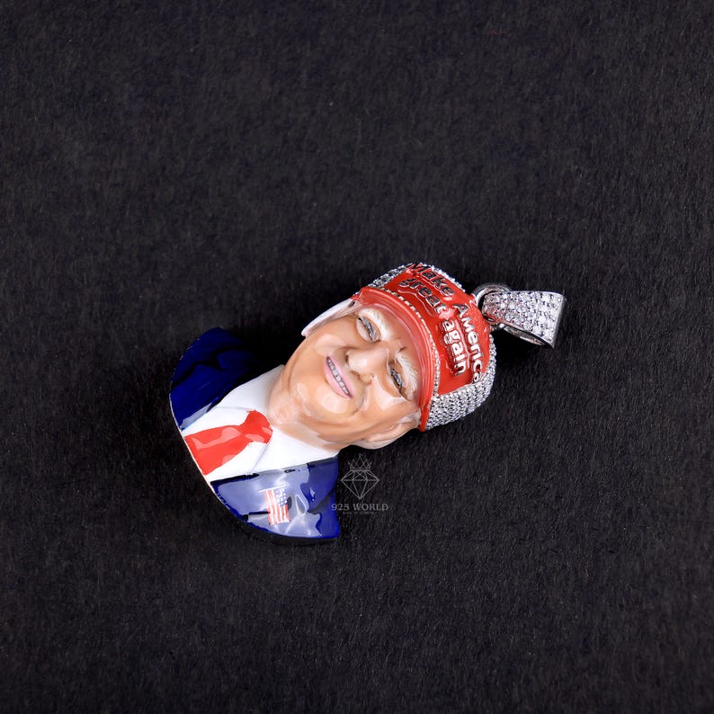 May include: A pendant featuring a smiling face with a red and white head covering that reads "Make America Great Again." The pendant has a silver bail and is set against a black background. The face is painted with realistic skin tones.