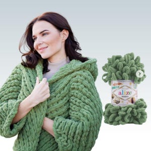 Pode incluir: Uma mulher vestindo um su&eacute;ter verde com capuz de malha. Ao lado dela est&aacute; uma bola de fio verde Alize Puffy. O su&eacute;ter &eacute; feito de malha grossa e texturizada. O fio &eacute; um estilo chenille macio e em la&ccedil;o.