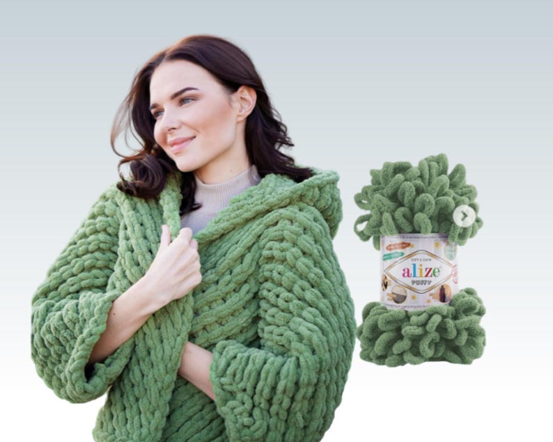 Pode incluir: Uma mulher vestindo um su&eacute;ter verde com capuz de malha. Ao lado dela est&aacute; uma bola de fio verde Alize Puffy. O su&eacute;ter &eacute; feito de malha grossa e texturizada. O fio &eacute; um estilo chenille macio e em la&ccedil;o.