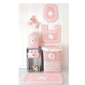 Pode incluir: Um conjunto de acess&oacute;rios de casa de banho em croch&eacute; rosa e branco, incluindo uma capa de assento de sanita, um tapete e um cesto de lixo. O conjunto apresenta detalhes de estrelas e flores, criando um visual coordenado e decorativo. Os acess&oacute;rios s&atilde;o exibidos contra uma parede branca.