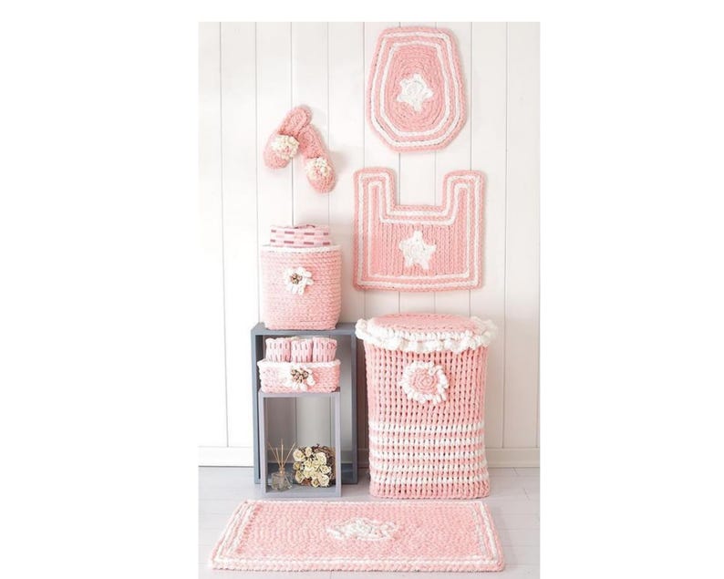 Pode incluir: Um conjunto de acess&oacute;rios de casa de banho em croch&eacute; rosa e branco, incluindo uma capa de assento de sanita, um tapete e um cesto de lixo. O conjunto apresenta detalhes de estrelas e flores, criando um visual coordenado e decorativo. Os acess&oacute;rios s&atilde;o exibidos contra uma parede branca.