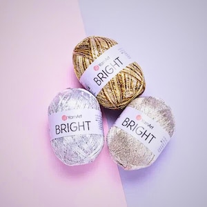 May include: Three skeins of metallic yarn in gold, silver, and champagne colors. Each skein has a white label with the word "BRIGHT" printed in black. The yarn is arranged on a pink and lavender background.