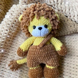 May include: A handmade crochet lion plush toy. The lion has a yellow body, brown mane, and brown dungarees. The toy has black eyes and a white muzzle. The lion is sitting on a cream-coloured blanket.