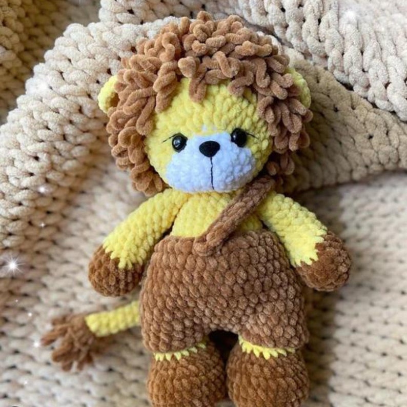 May include: A handmade crochet lion plush toy. The lion has a yellow body, brown mane, and brown dungarees. The toy has black eyes and a white muzzle. The lion is sitting on a cream-coloured blanket.