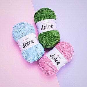 May include: Three balls of soft yarn in pastel shades: light blue, green, and pink. Each ball has a white label with the word "dolce" printed in black. The yarn is displayed against a pink and purple backdrop.