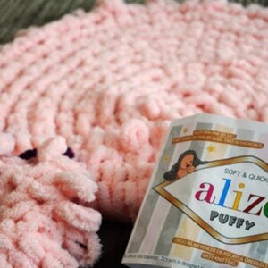 Pode incluir: Um close-up de um cobertor de malha rosa macio e fio. A embalagem do fio diz "alize PUFFY" com um urso de desenho animado. O cobertor tem um design texturizado e em la&ccedil;o, e o fio parece fofo e convidativo.