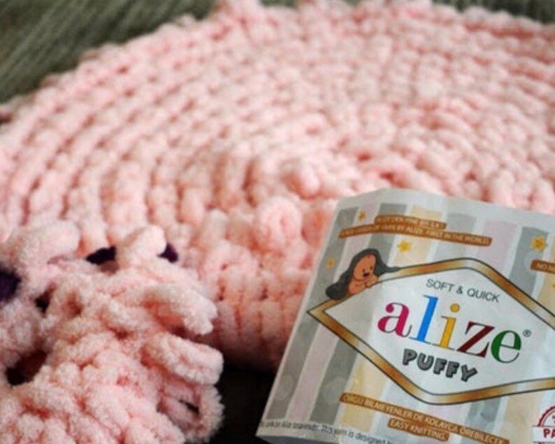 Pode incluir: Um close-up de um cobertor de malha rosa macio e fio. A embalagem do fio diz "alize PUFFY" com um urso de desenho animado. O cobertor tem um design texturizado e em la&ccedil;o, e o fio parece fofo e convidativo.