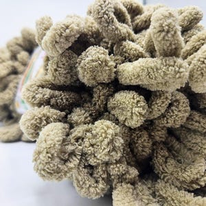 Pode incluir: Close-up de uma bola de l&atilde; macia e texturizada em marrom claro. A l&atilde; tem uma apar&ecirc;ncia em la&ccedil;o, semelhante a um pompom, criando uma textura macia e convidativa. Ideal para projetos de tric&ocirc; ou croch&ecirc;.