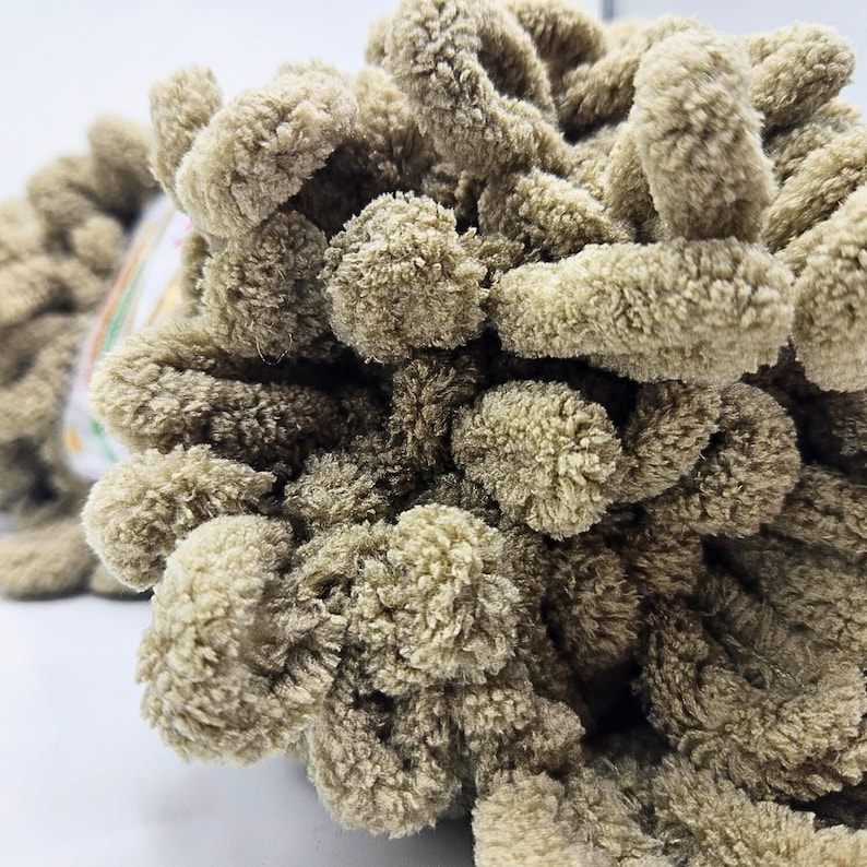 Pode incluir: Close-up de uma bola de l&atilde; macia e texturizada em marrom claro. A l&atilde; tem uma apar&ecirc;ncia em la&ccedil;o, semelhante a um pompom, criando uma textura macia e convidativa. Ideal para projetos de tric&ocirc; ou croch&ecirc;.