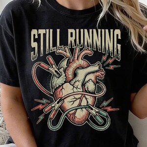 Op de afbeelding: Een zwart t-shirt met korte mouwen en een afbeelding van een anatomisch hart verbonden met startkabels. De tekst "STILL RUNNING" staat in een verweerd wit lettertype boven de hartillustratie, die rode en groene kabelaccenten bevat.