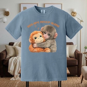 Puede incluir: Camiseta azul claro con un gráfico de dos monos abrazándose. El texto naranja sobre los monos dice "you are never alone" con un pequeño símbolo de corazón. Los monos son marrones y grises.
