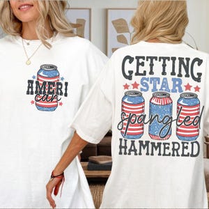 May include: A white short-sleeve t-shirt featuring a graphic of a beverage can with red and white stripes and blue stars. The front displays the text AMERI-CAN. The back features three beverage cans and the text GETTING STAR SPANGLED HAMMERED.